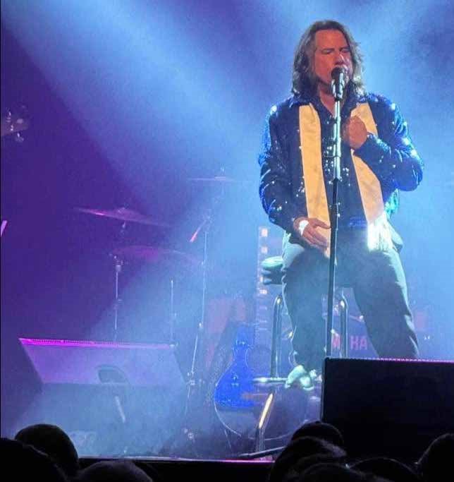 man with long brown hair singing into a microphone with a yellow scarf on